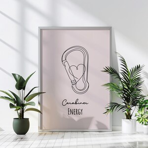 May include: A framed print featuring a minimalist line drawing of a carabiner with a heart inside. The words "Carabiner ENERGY" are written in a script font below the drawing. The background is a soft pink color.
