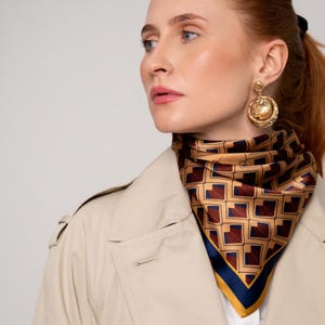 May include: A patterned silk scarf in tan, navy, and burgundy, worn with a beige trench coat and gold earrings. The scarf features a repeating geometric design. The scarf is a square shape and is worn around the neck.