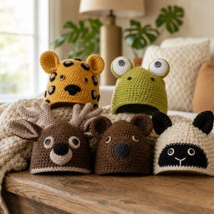 May include: Five crocheted animal hats featuring a yellow leopard with spots, a green frog with button eyes, a brown deer with antlers, a brown bear with rounded ears, and a cream-coloured sheep with black ears and a black face patch.