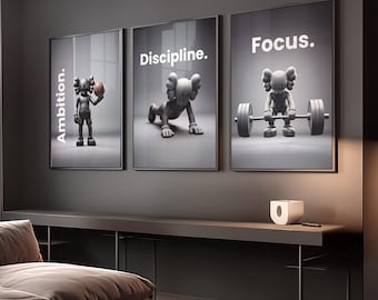 May include: Three framed art prints with a gray and white color scheme. Each print features a cartoon figure in different poses with the words "Ambition," "Discipline," and "Focus." The prints are displayed above a dark gray console table.