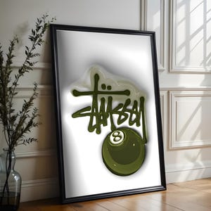 May include: A framed art print featuring the word "STUSSY" in a green, stylized font, with an olive green eight-ball graphic. The artwork is set against a white background and framed in black.