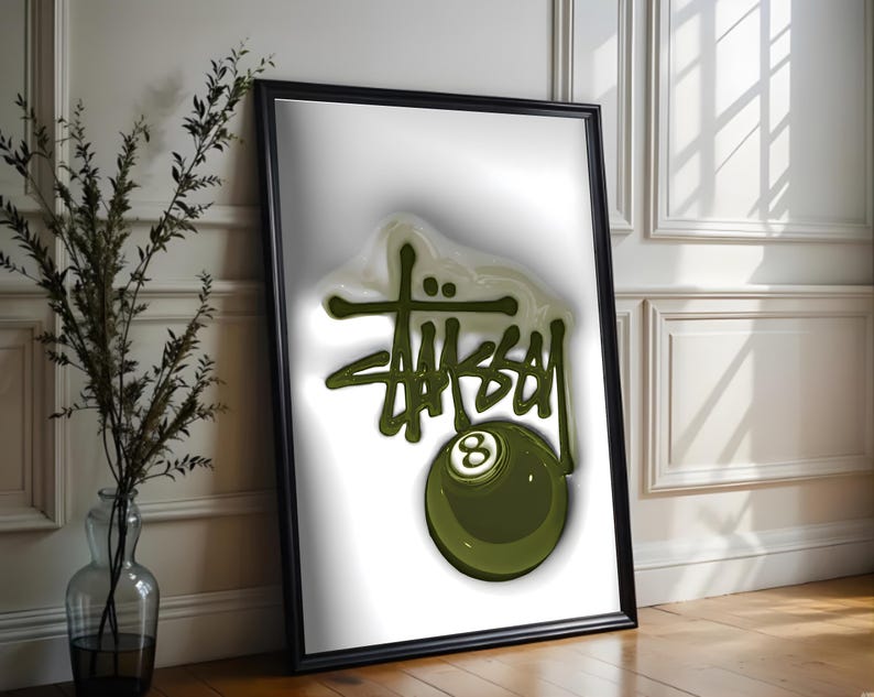 May include: A framed art print featuring the word "STUSSY" in a green, stylized font, with an olive green eight-ball graphic. The artwork is set against a white background and framed in black.