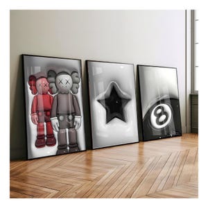 May include: Three framed art prints leaning against a wall. The prints feature pop art designs: two cartoon figures, a black star, and a black and white eight ball. The frames are black, and the artwork is displayed on a wooden floor.
