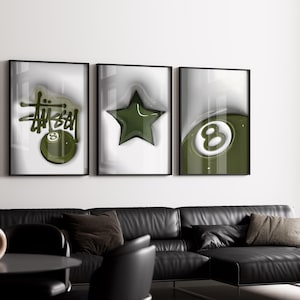 May include: Three framed art prints with black frames. The prints feature a green and white color scheme with a Stussy logo, a star, and an eight ball design. The art is displayed above a black leather sofa.