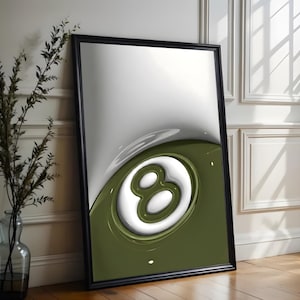 May include: Framed art print featuring a stylized 8-ball design. The artwork has a glossy finish with a white and olive green color scheme. The print is in a black frame and leans against a white wall.