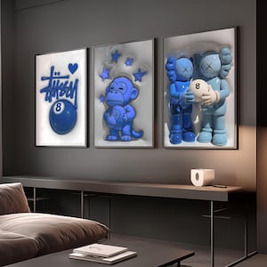 Op de afbeelding: Drie ingelijste wandkunstwerken met blauw-witte ontwerpen. Eén stuk toont het woord "Stussy" met een hart en een blauwe 8-ball. Een ander toont een blauwe aap die een kleinere aap vasthoudt. De derde toont twee blauwe figuren die een witte bal met het nummer 8 vasthouden.