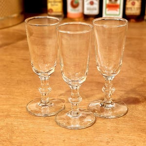 May include: Three clear, stemmed shot glasses with a classic design. The glasses have a round base, a decorative stem, and a slightly flared top. They are arranged on a wooden surface, with miniature liquor bottles in the background.