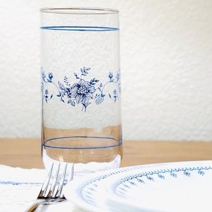 May include: A clear glass with blue floral design and a blue rim, a white plate with a blue floral border, and a silver fork rest on a white lace tablecloth. The glass and plate are on a wooden surface.