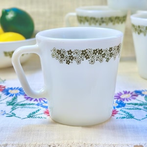 May include: White ceramic mugs with a floral design in shades of green and brown. The mugs have a simple handle and a slightly flared shape. A bowl of lemons and limes is in the background. The mugs are on a floral embroidered tablecloth.