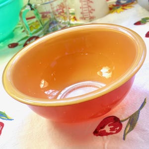 May include: An orange and yellow glass mixing bowl. The bowl has a rounded shape with a smooth, glossy finish. The rim of the bowl is a lighter shade of yellow, creating a subtle contrast. The bowl is sitting on a floral patterned tablecloth.