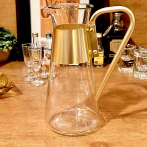 May include: A clear glass pitcher with a gold-colored metal handle and a gold band around the middle. The pitcher has a spout for easy pouring. The design is simple and elegant, suitable for serving beverages.