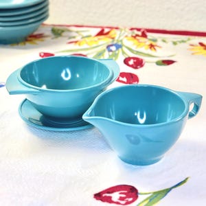 May include: A collection of turquoise-colored serving bowls and a small pitcher. The bowls have a rounded shape with handles, and one sits on a matching saucer. The pitcher has a spout and a handle. The items are on a floral patterned tablecloth.