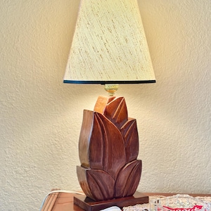 May include: A table lamp with a dark brown, carved wooden base shaped like layered flower petals. The lamp features a conical, textured off-white lampshade with a thin black trim at the top and bottom edges, resting on a rectangular wooden base.