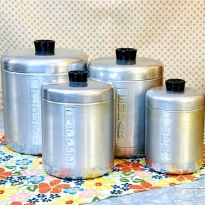 May include: A set of four cylindrical brushed aluminum canisters with black knobs on the lids. Each canister features embossed vertical text identifying the contents as FLOUR, COFFEE, SUGAR, and TEA, arranged in descending order of size.
