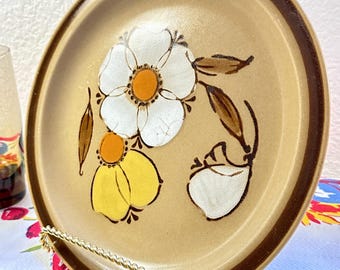 Puede incluir: Un plato redondo de cerámica con base color canela y borde marrón oscuro. El centro presenta un diseño floral pintado con una flor blanca grande con centro naranja, una flor amarilla más pequeña y un capullo blanco, con hojas y tallos marrones.