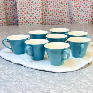 May include: A set of seven ceramic cups featuring a vibrant teal exterior and a smooth cream-colored interior. Each cup has a curved handle and a wide, flared rim, arranged in a cluster on a white fabric surface.