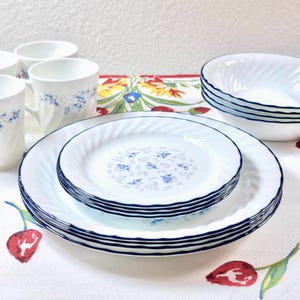 May include: A set of white dinnerware featuring a blue floral pattern and a dark blue rim. The collection includes stacked dinner plates, smaller salad plates, bowls, and mugs, all characterized by a subtle spiral texture on the surface.