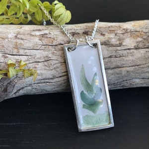 Puede incluir: Un collar de cadena de plata con un colgante rectangular que presenta un diseño de hoja verde estilo acuarela sobre un fondo blanco.