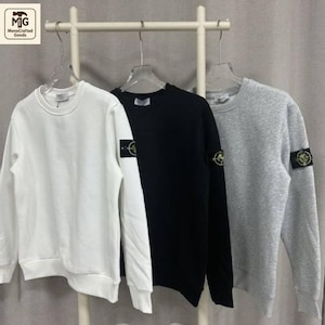 Könnte beinhalten: Drei Sweatshirts mit Rundhalsausschnitt in Weiß, Schwarz und Grau, die an einem Gestell hängen. Jedes Sweatshirt hat einen Aufnäher am linken Ärmel. Die Sweatshirts sind aus einem weichen Material. Das Logo "MonoCrafted Goods" befindet sich oben links.