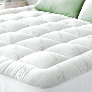 May include: A white quilted mattress topper with a diamond pattern. The topper has the word "Bamboo" printed on it in a light gray color. Two green pillows are visible in the background.
