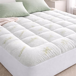 May include: White quilted mattress topper with a bamboo leaf pattern. Two light green pillows rest on the bed. The bed is made up and ready for use. The topper has a fitted skirt.
