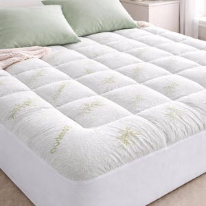 May include: White quilted mattress topper with a bamboo leaf pattern. Two light green pillows rest on the bed. The bed is made up and ready for use. The topper has a fitted skirt.
