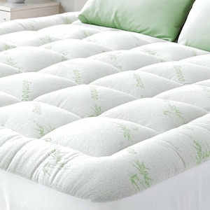 May include: White mattress topper with a quilted design. The topper has a textured surface and is printed with the word "Bamboo" and bamboo leaf illustrations in light green. Two green pillows are visible in the background.