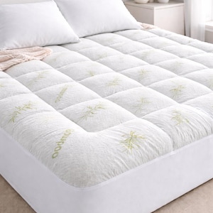 May include: A white bamboo mattress topper with a quilted design and green bamboo leaf pattern. The topper is on a bed with white pillows and a white bed skirt. The bed is in a well-lit room.