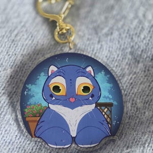 Puede incluir: Un llavero azul con temática de gato con un cierre dorado en forma de corazón. La ilustración del gato tiene ojos amarillos grandes y una lengua rosa. El fondo presenta una escena nocturna con una pequeña maceta.
