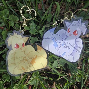 May include: Two acrylic keychains featuring cartoon fox-like creatures with red eyes. One is yellow with an orange tail, and the other is white with blue accents. Both have gold-colored heart-shaped clasps and are set against a backdrop of green grass.