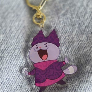May include: A clear acrylic keychain featuring a cartoon cat character in shades of purple and pink. The cat has an open mouth and is wearing a patterned outfit. The keychain has a gold-colored heart-shaped clasp.