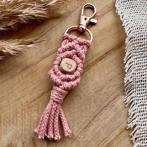 Könnte beinhalten: Handgefertigter Makramee-Schlüsselanhänger in Staubrosa. Der Schlüsselanhänger hat eine Holzperle mit dem Buchstaben "P" und eine Quaste. Er hat einen roségoldenen Verschluss zum Befestigen an Schlüsseln oder einer Tasche.
