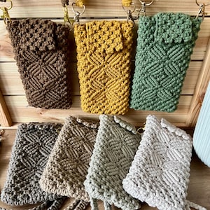 Könnte beinhalten: Eine Sammlung von Makramee-Handytaschen in verschiedenen Farben, darunter Braun, Gelb und Grün, die an einer Holzkonstruktion hängen. Die Taschen weisen ein geometrisches Muster auf und werden mit ihren Trägern und Metallverschlüssen präsentiert. Zusätzliche Taschen in Grau und Beige sind ebenfalls zu sehen.