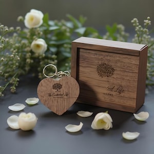 Può includere: Un portachiavi a forma di cuore in legno con anello dorato, inciso con logo ad albero e testo. Accanto, una scatola di legno con logo ad albero simile e le parole "red Life" e "NAPHY". Petali e fiori bianchi sparsi.