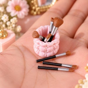 Peut inclure: Un porte-pinceaux miniature rose clair matelassé avec de petites perles blanches, contenant plusieurs minuscules pinceaux de maquillage à poils bruns et viroles argentées, avec trois pinceaux supplémentaires posés à plat.