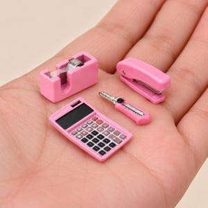 Peut inclure: Un ensemble miniature de fournitures de bureau rose comprenant un dévidoir de ruban adhésif, une agrafeuse, un cutter et une calculatrice avec un écran noir et des touches noires, disposés ensemble sur une surface plane.