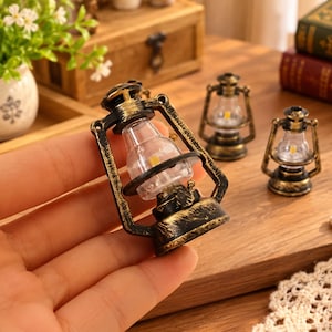 Può includere: Una lanterna in miniatura in stile olio con finitura nera e dorata anticata, un globo in plastica trasparente e un piccolo componente luminoso giallo all'interno. La lanterna presenta una struttura effetto metallo con manico.