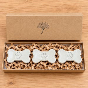 May include: Three light gray ceramic bone-shaped ornaments, each featuring an embossed paw print, are nestled in a rectangular brown cardboard gift box filled with shredded paper. The box lid displays a black line drawing of a ginkgo leaf.