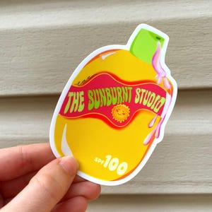 May include: A yellow sticker shaped like a sunscreen bottle with a green cap. The sticker features the text "THE SUNBURNT STUDIO" in red and a small sun graphic. It also displays "SPF 100".