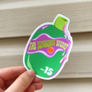 May include: A green and purple sticker in the shape of a sunscreen bottle. The sticker features the text "THE SUNBURNT STUDIO" and "SPF 15". The design includes a sun graphic and pink dripping accents.