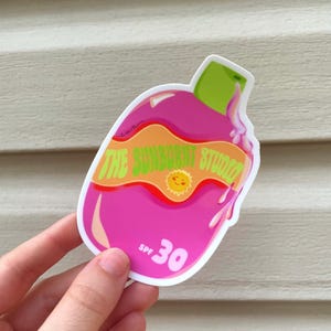May include: A pink sticker in the shape of a sunscreen bottle with the text "THE SUNBURNT STUDIO" and "SPF 30". The sticker has a green cap and orange accents, with a smiling sun graphic.
