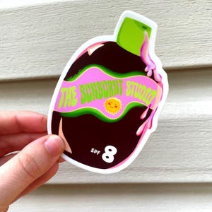 May include: A sticker depicting a sunscreen bottle design. The bottle is mainly dark brown with a pink label that reads "THE SUNBURNT STUDIO" and a small sun graphic. The sticker also includes "SPF 8" in white.