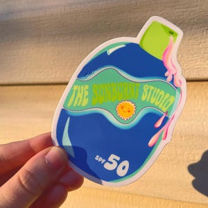 May include: A sticker featuring a blue bottle design with the text "THE SUNBURNT STUDIO" and "SPF 50". The sticker has a green cap and pink dripping accents, with a smiling sun graphic.