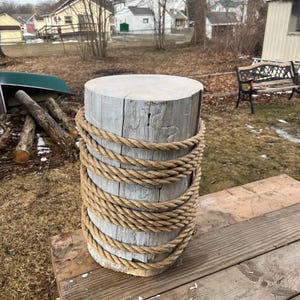 May include: A cylindrical wooden post, painted light grey, wrapped with thick, natural-coloured rope. The post sits on a wooden surface. The rope is tightly wound around the post, creating a rustic, nautical aesthetic. The post is likely used for decorative purposes.
