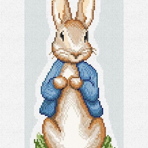 May include: A brown rabbit wearing a blue jacket stands in a patch of green grass. The rabbit has long ears and a white nose.