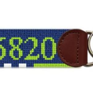 May include: A navy blue key fob with the number "06820" stitched in lime green. It features a brown leather backing and silver key rings.