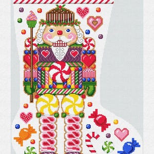 Puede incluir: Un calcetín navideño blanco con un diseño colorido de cascanueces. El calcetín está adornado con caramelos, corazones y el nombre "Adrianna" en verde. El diseño incluye detalles en rojo, amarillo, verde y azul.