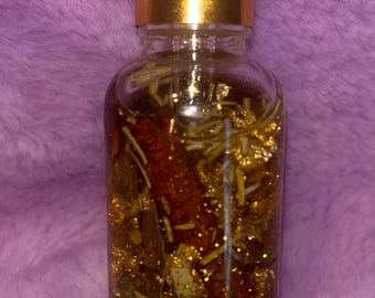 May include: A clear glass bottle with a gold-colored cap and white dropper. The bottle is filled with a golden liquid and contains herbs and gold flakes. The background is a fuzzy purple surface.