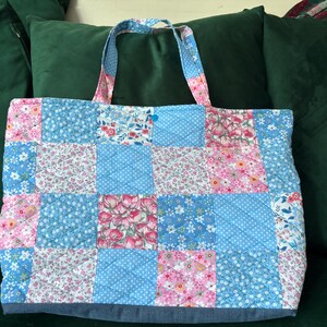 Peut inclure: Un sac fourre-tout matelassé avec un motif patchwork. Le sac présente des carrés de tissu bleu clair, rose et à motifs floraux. Les poignées et le bas sont bleus unis. Le sac est posé sur une surface verte.