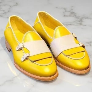 May include: A pair of bright yellow leather loafers featuring a wide off-white strap across the vamp. Each shoe is secured with two polished silver-tone metal buckles on the side and sits on a light tan wooden sole with a low heel profile.
